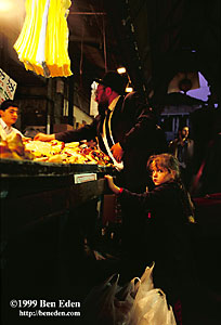 A little girl waits next to the many shopping bags while her father, an orthodox Jew wearing black suit with a tie and a hat, pays at a bakery stand at the Shuk Ha'Carmel market in Jerusalem, Israel