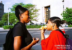 Two black teenage girls witch carefully styled hairdos stand across the street from the Brooklyn Public Library and eat hot dogs in New York City, USA