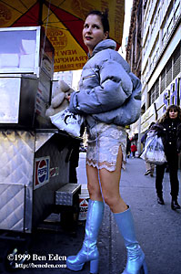 A young woman wearing blue-silver platform boots and a window-lace mini-skirt holds a teddybear next to a hot dog stand in Manhattan, New York City