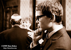 Czech Jewish boy wearing black glasses, carefully groomed and formally dressed holds his fist to his chin as he contemplates the dancing scene at a Prague Chanukah (Hanukah) celebration. An older couple dances in the background.