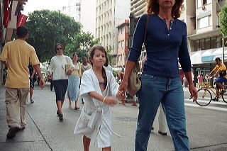 A little blonde girl wearing a white bathrobe over bikini smirks as she's being towed by her mother on a busy Rio de Janeiro street
