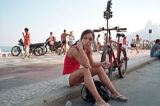 A young brazilian woman in white shorts and a red tanktop sits on the sidewalk next to her bicycle and watches the passers-by on Ipanema beach, Rio de Janeiro
