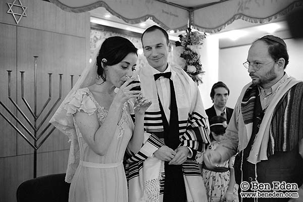 The bride drinks wine under the Chuppah watched by the groom and rabbi